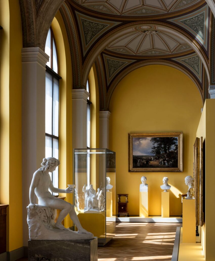 A sunlit gallery with classical sculptures and paintings in a museum with ornate arched ceilings and yellow walls.