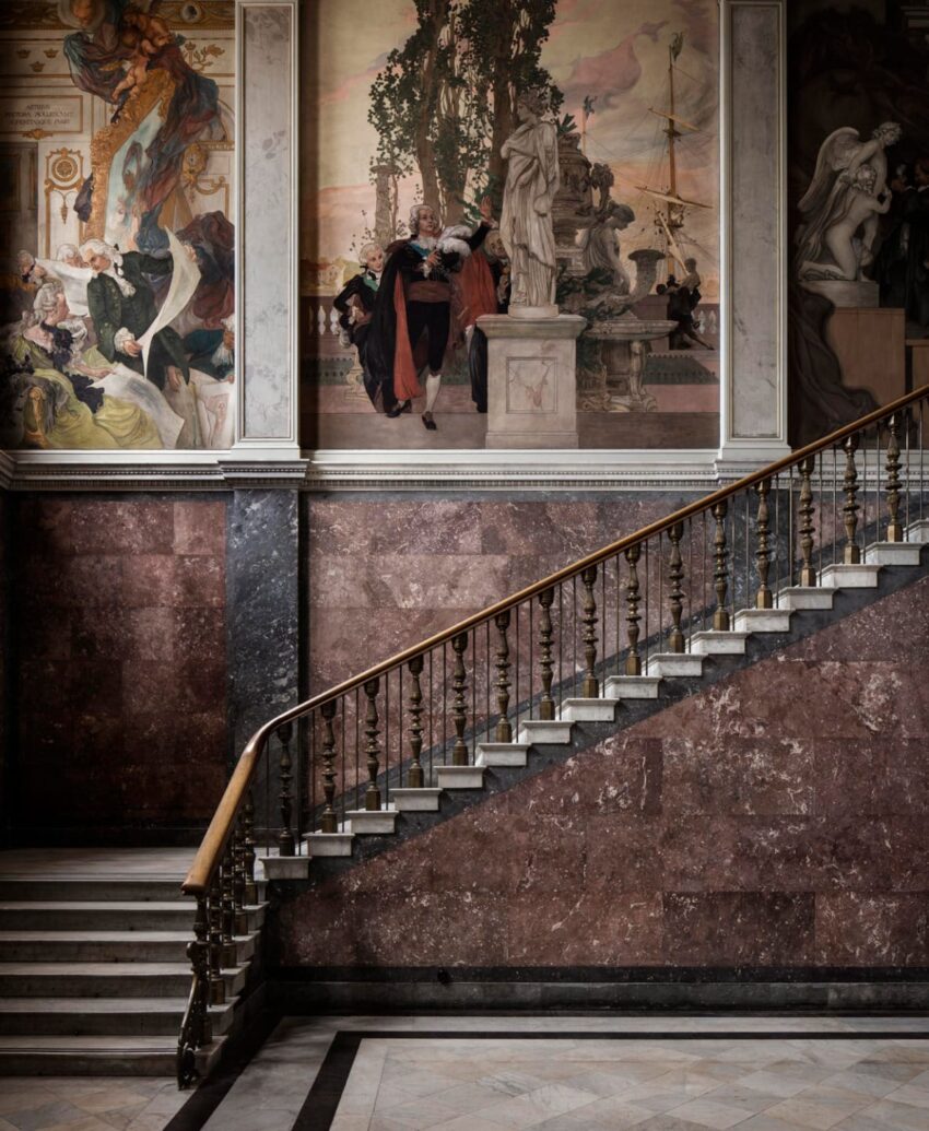 Elegant staircase with ornate railing leading to large, detailed wall murals in a historic building interior.