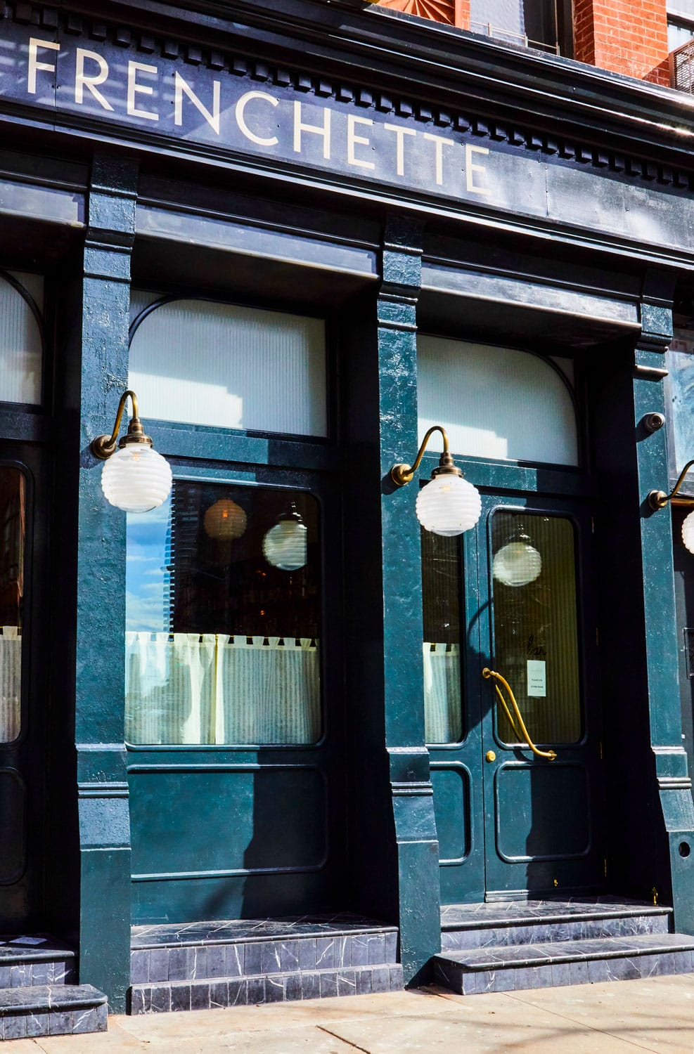 Exterior of Frenchette restaurant with blue doors, circular light fixtures, and brick wall background.