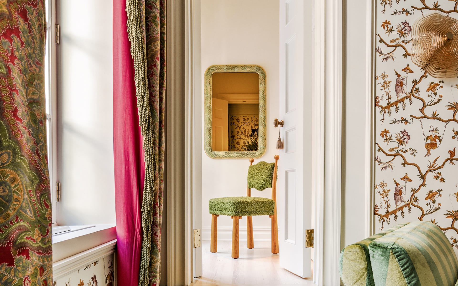 Elegant room with patterned wallpaper, colorful drapes, green chair, and decorative gold-framed mirror next to open door.