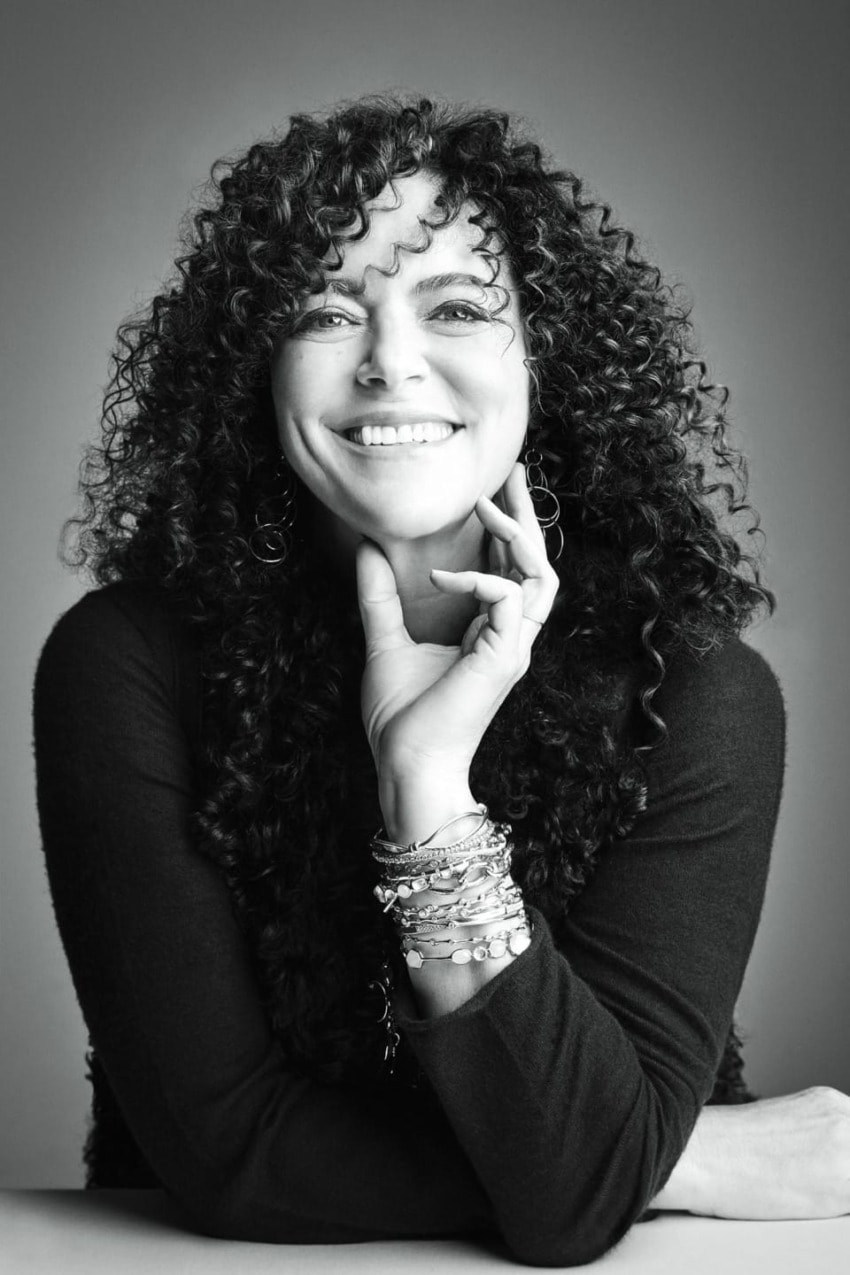 Person with curly hair smiling, resting chin on hand, wearing a dark sweater and bracelets.