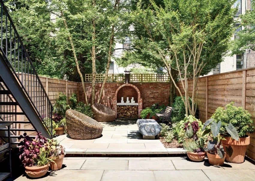 Lush backyard garden with trees, potted plants, cozy seating, and a brick firewood storage area on a sunny day.