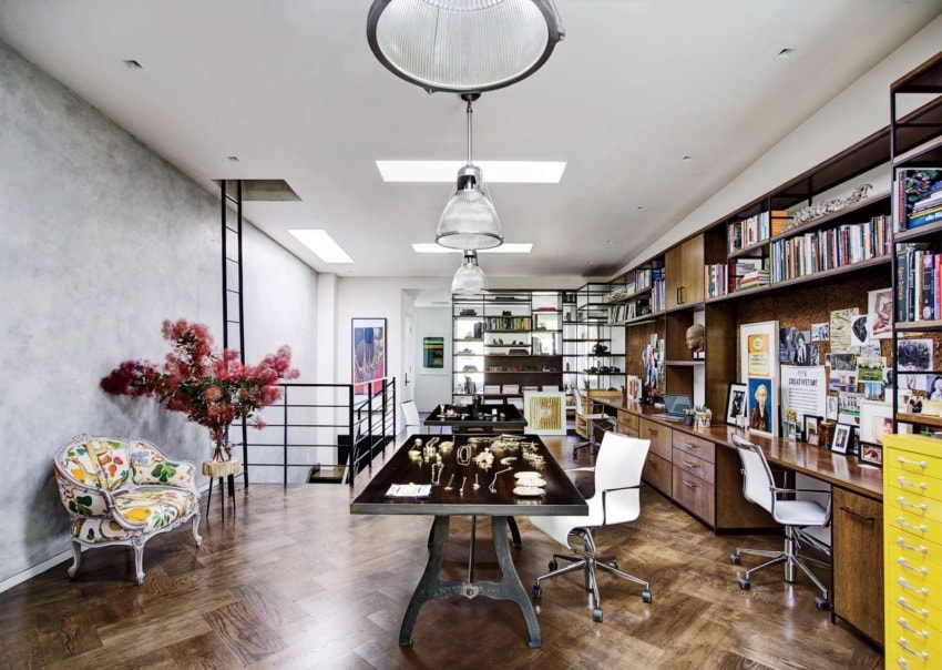 Modern home office with wooden floors, artwork, bookshelves, and a large table with decorative pieces, featuring ample natural light.