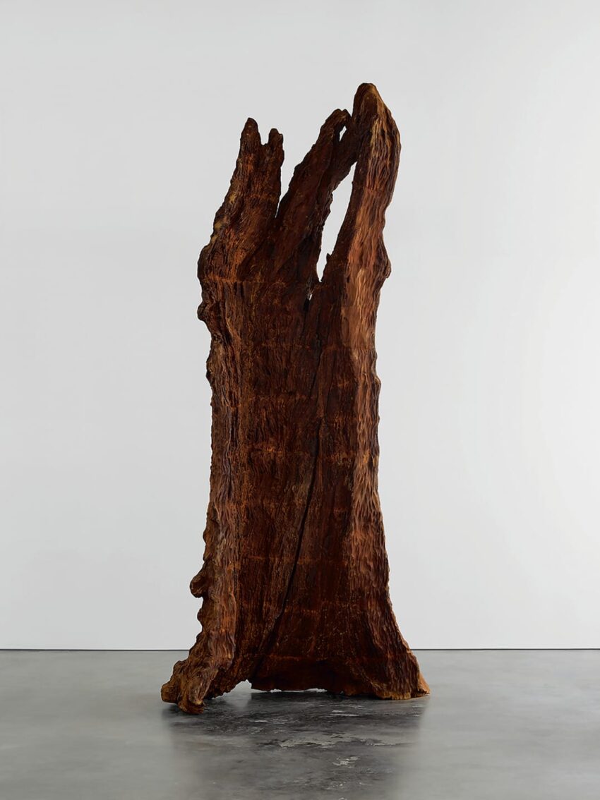 Large wooden sculpture resembling a tree trunk displayed in a minimalist gallery setting with a concrete floor.