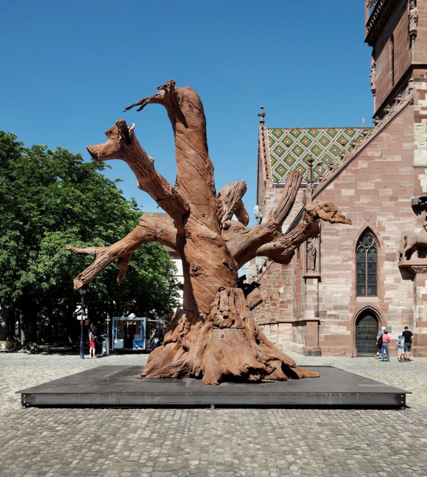 Large wooden tree sculpture in an urban square with a historic building and trees in the background.