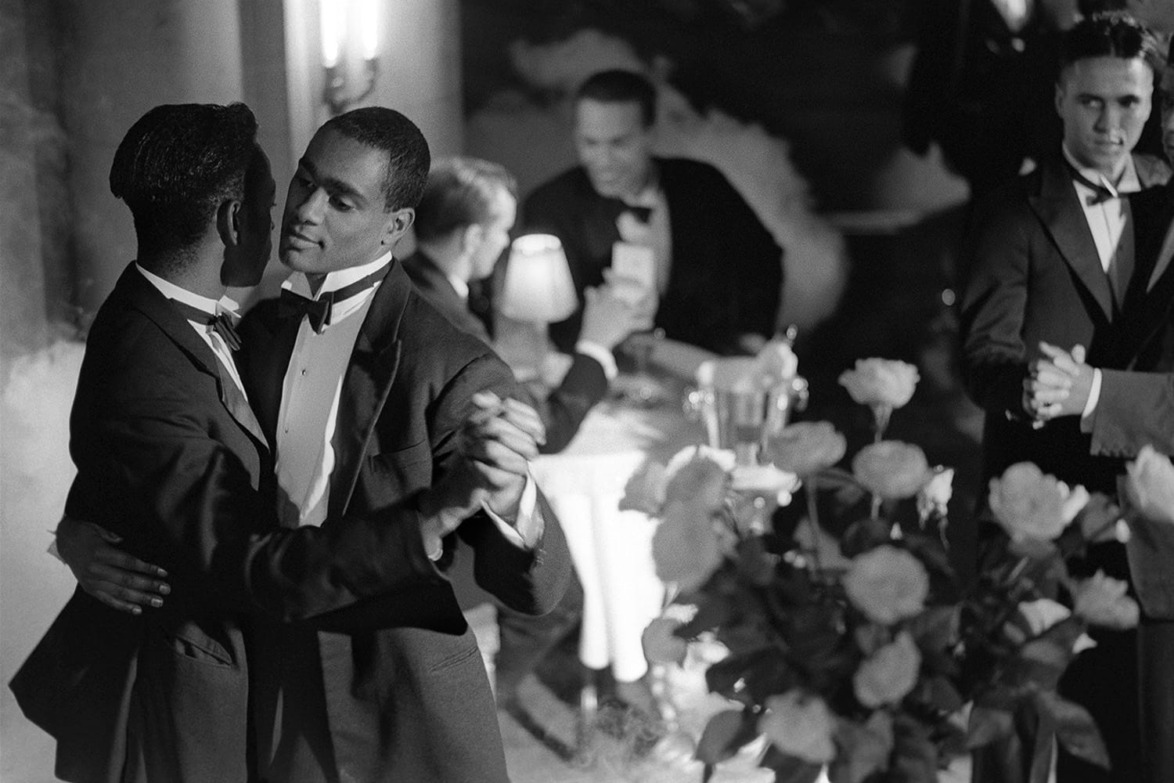 Two men in tuxedos dancing in a dimly lit, elegant setting with roses and people seated at tables in the background.