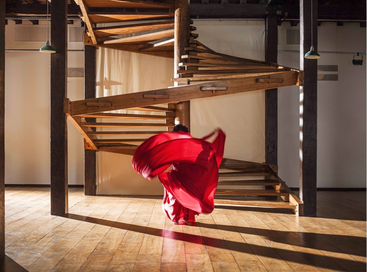 Person in a flowing red dress dances gracefully under a wooden spiral staircase in a sunlit room.