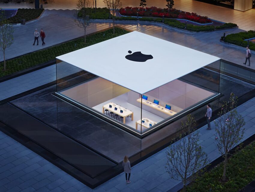 Apple store with glass architecture and large logo, showcasing modern design and visible tech products inside.