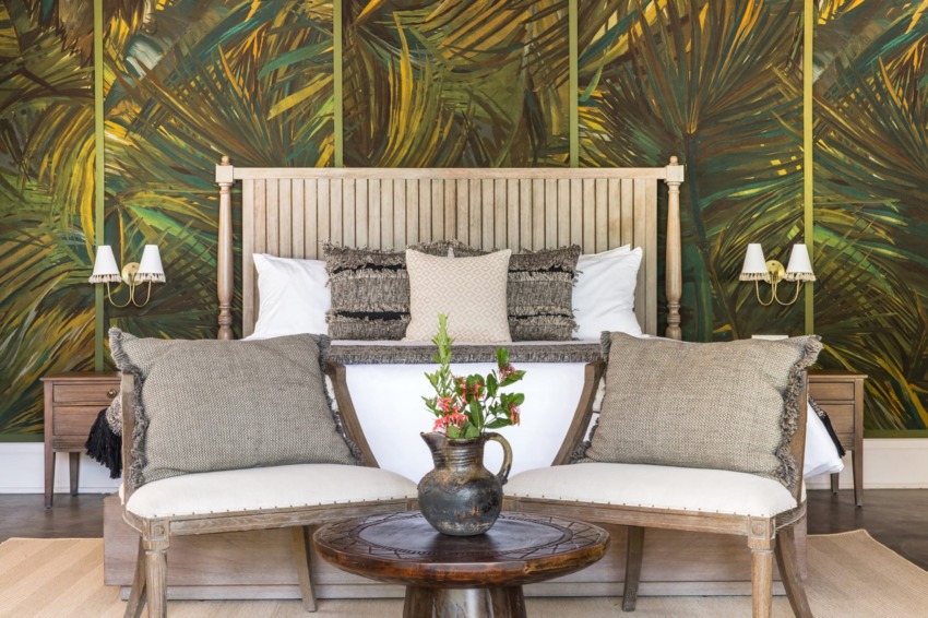 Tropical-themed bedroom with wooden bed, decorative pillows, two chairs, and a small table with a vase on it.