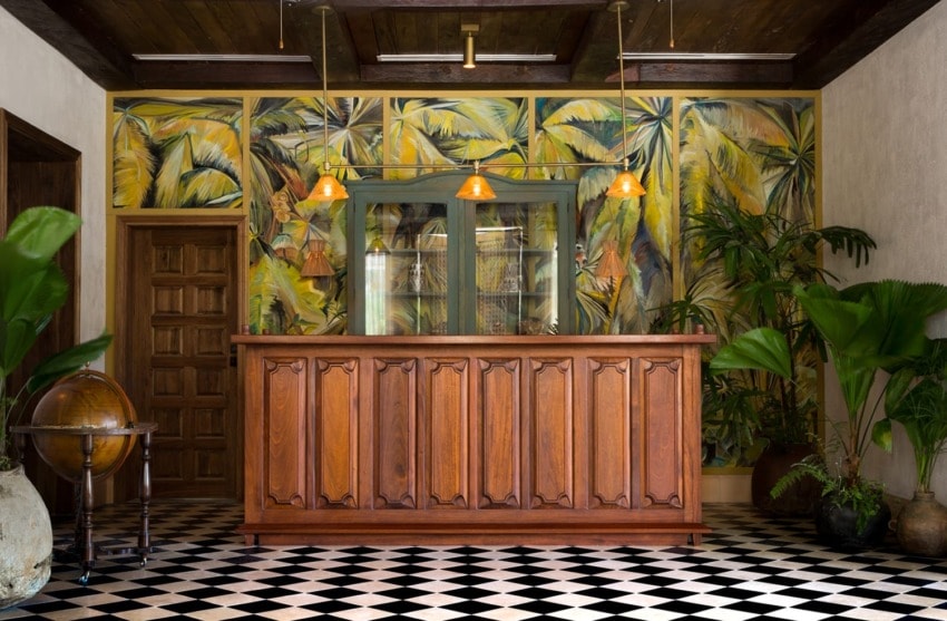 Vintage hotel lobby with wooden reception desk, tropical mural, globe, and checkered floor tiles.