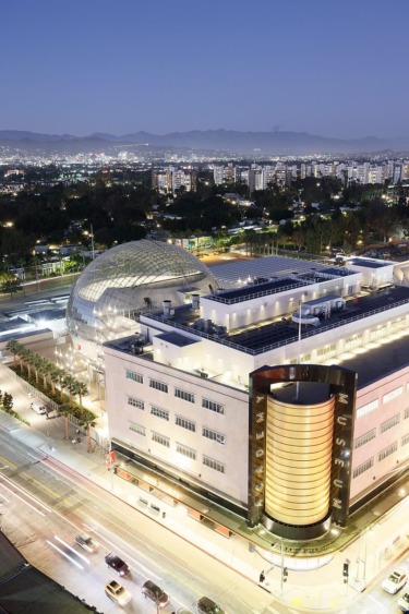 Aerial view of a modern cityscape at dusk featuring an illuminated dome and adjacent building with city skyline in background.