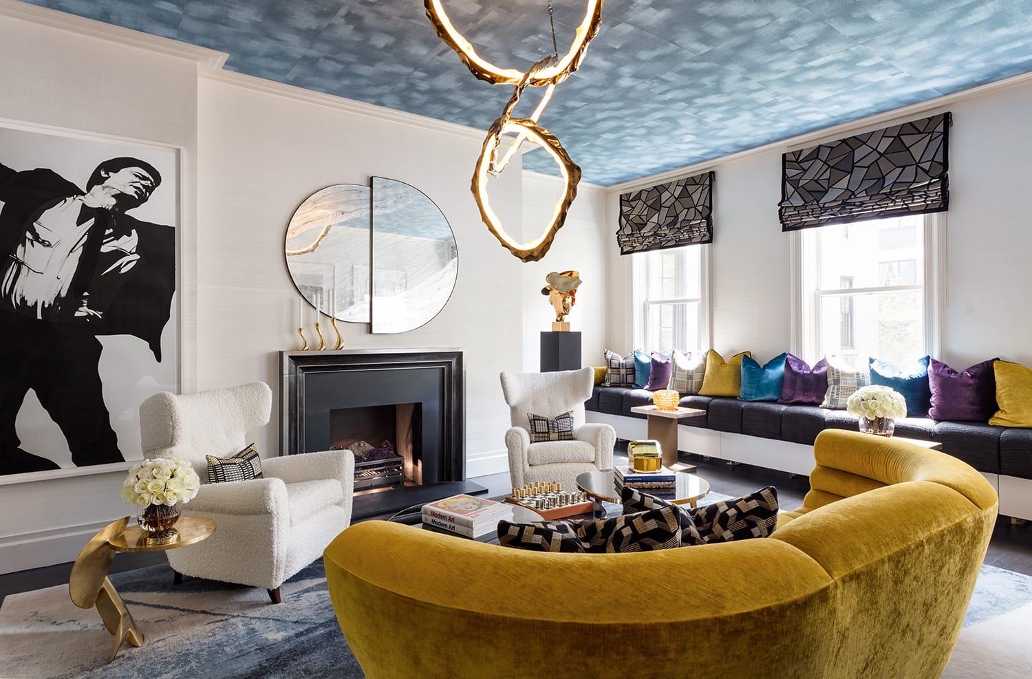 Modern living room with yellow sofa, abstract chandelier, large artwork, and cozy fireplace with blue ceiling accents.