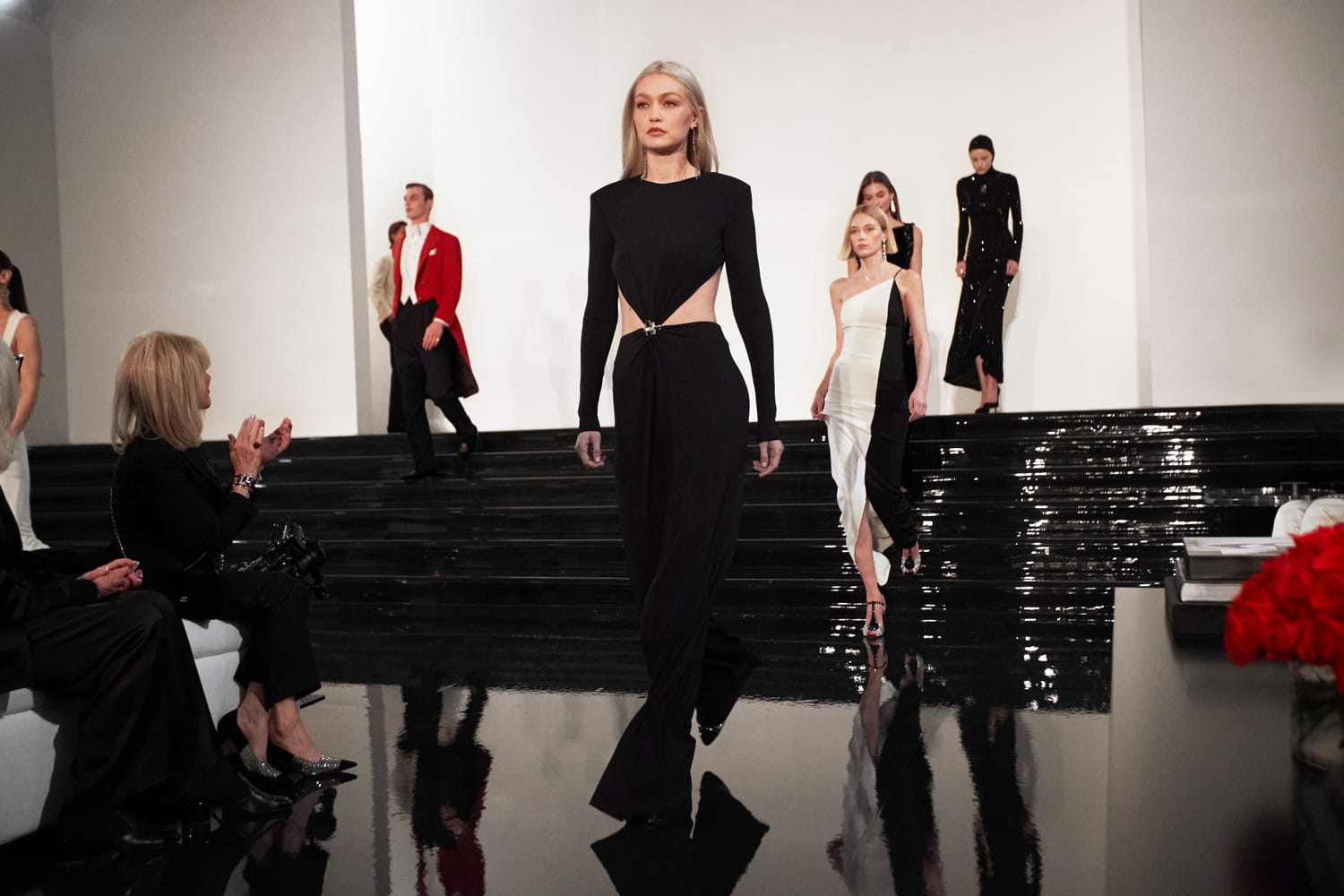 Models walking on a shiny black runway showcasing elegant black and white fashion outfits, audience clapping.