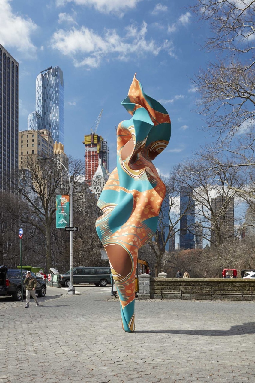 Colorful abstract sculpture in a city park with tall buildings and trees in the background under a blue sky.