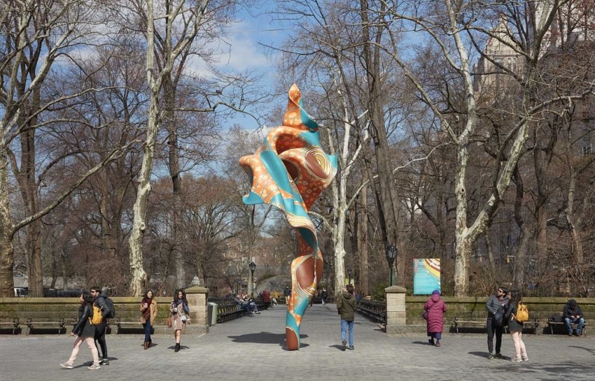 Colorful abstract sculpture in a park with bare trees, surrounded by people walking on a cloudy day.