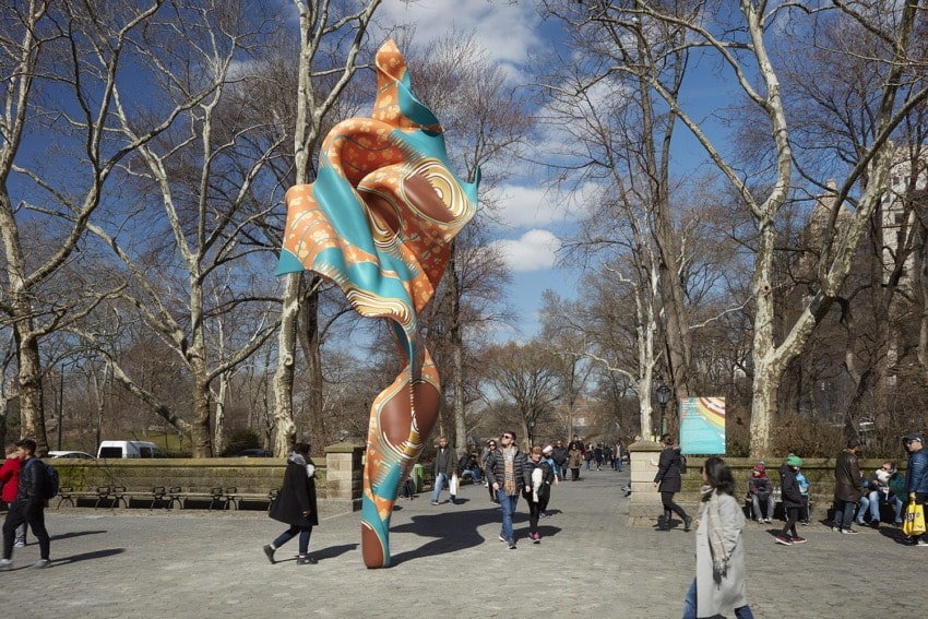 Colorful abstract sculpture stands tall in a park surrounded by people walking on a sunny day in early spring.