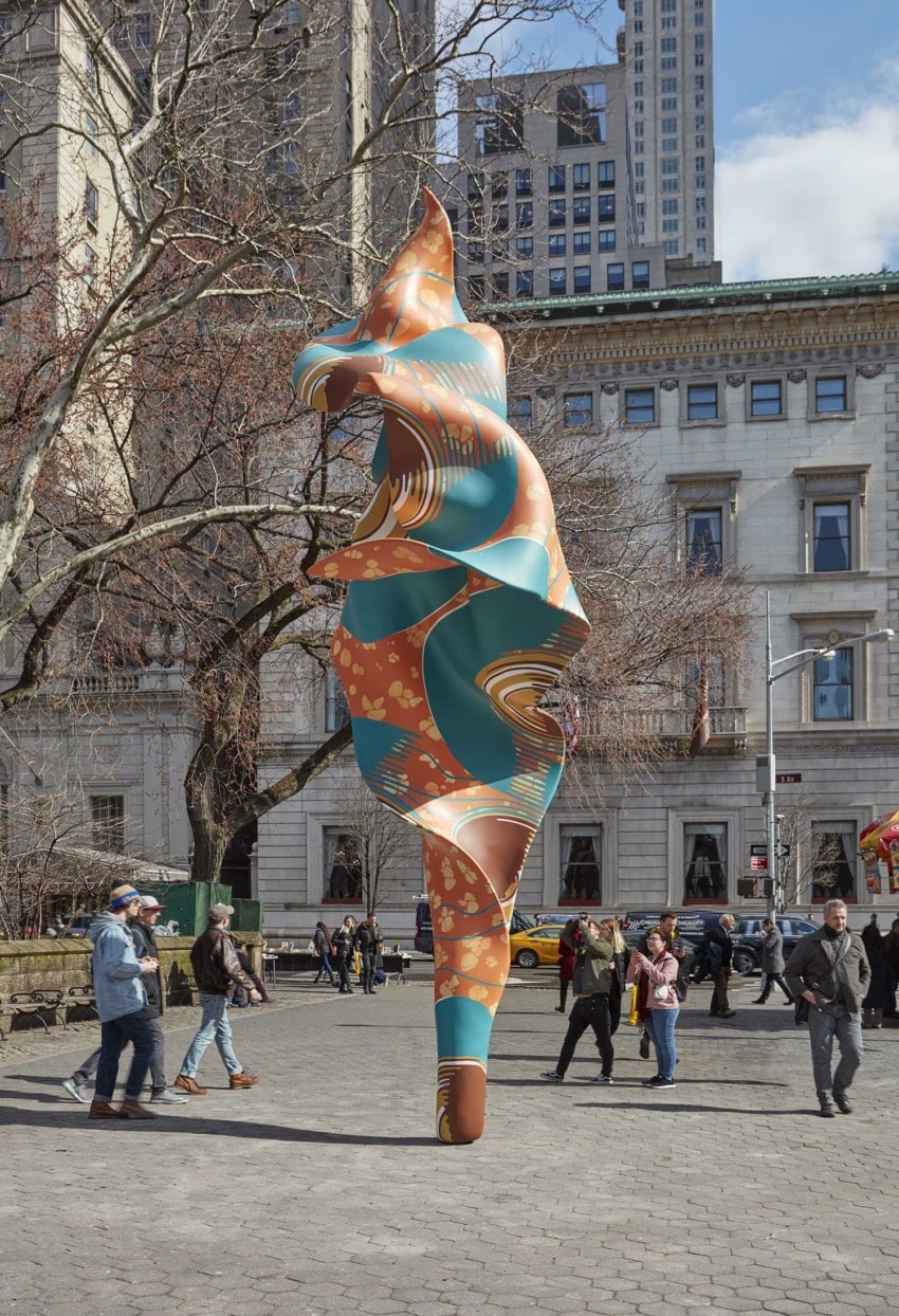 Colorful abstract sculpture in urban park setting surrounded by walking people and trees, with buildings in background.