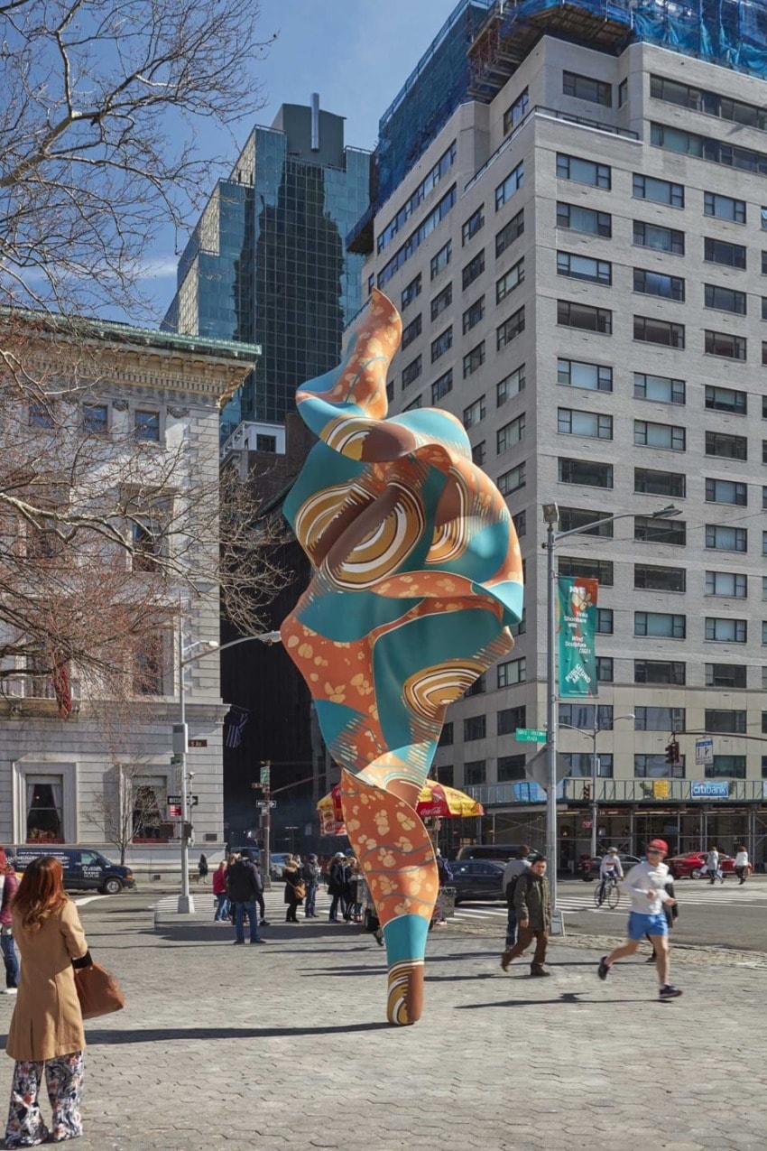 Colorful abstract sculpture in an urban plaza with people walking, tall buildings, and trees in the background.