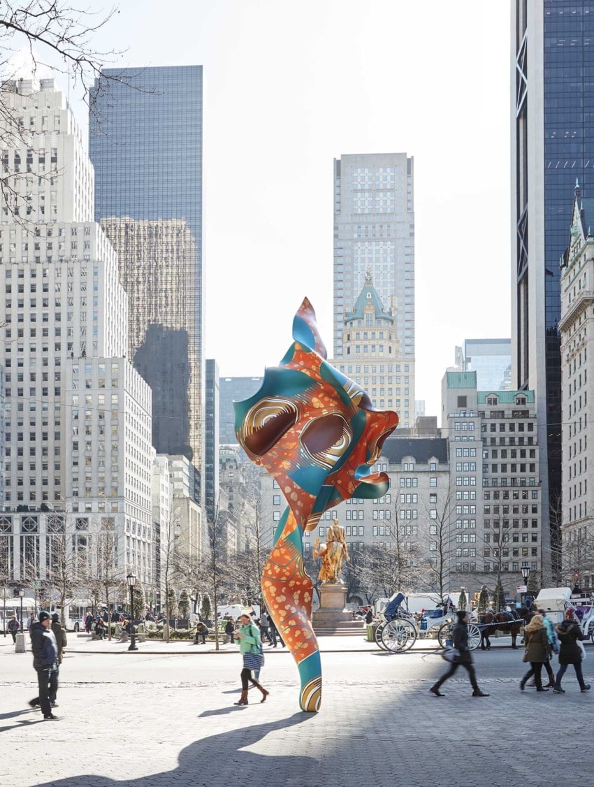Colorful abstract sculpture in a bustling city plaza with skyscrapers in the background and people walking around.
