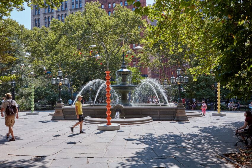 Circular fountain in a park with surrounding trees, people walking nearby, and buildings in the background on a sunny day.