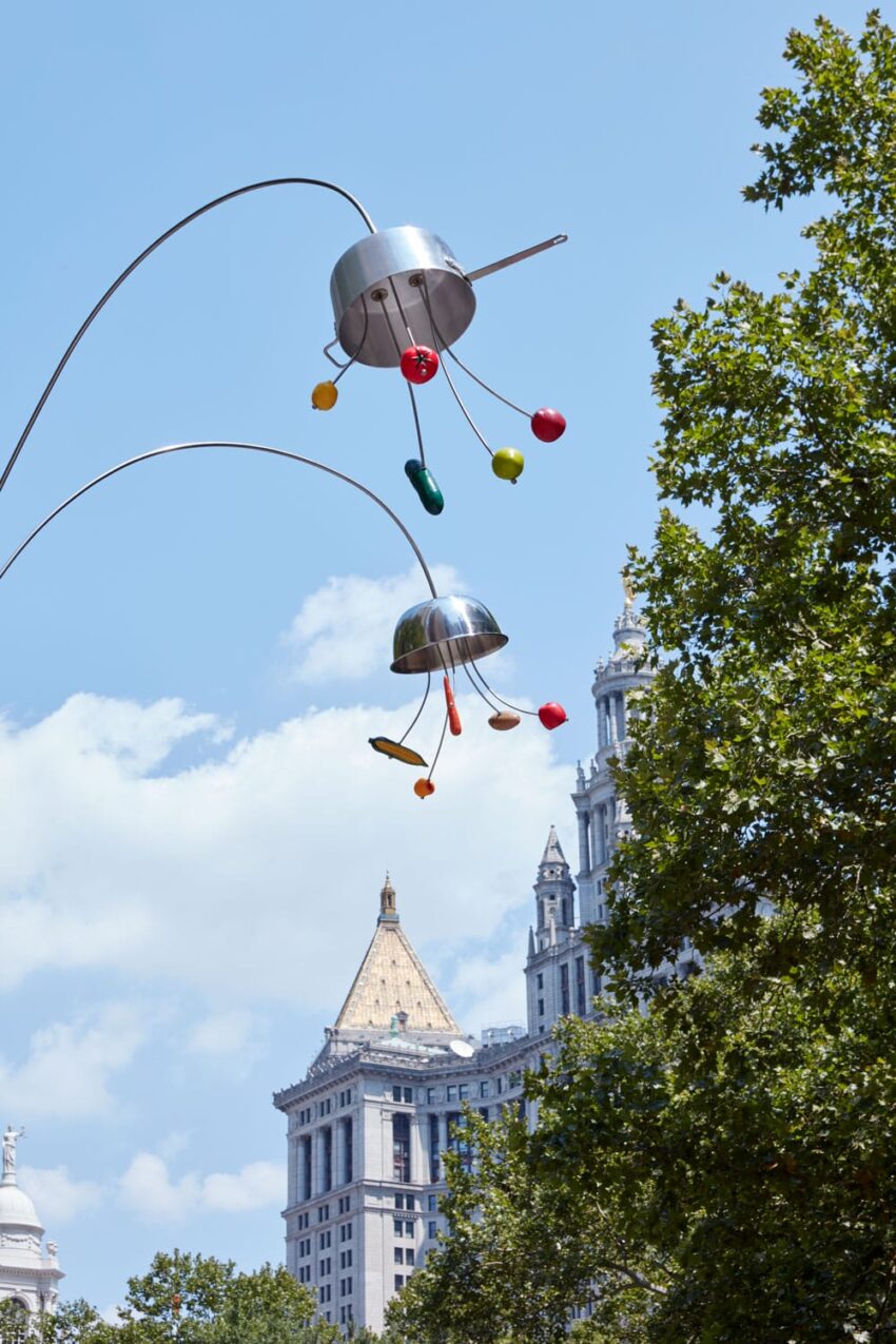 Colorful abstract sculpture with metal rods and shapes against a blue sky and historic building backdrop.