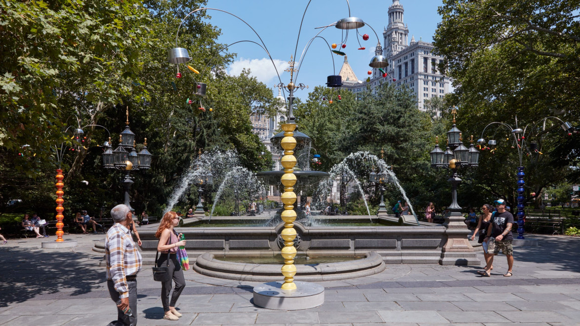 People walking near a decorative fountain in a park with trees and a historic building in the background.