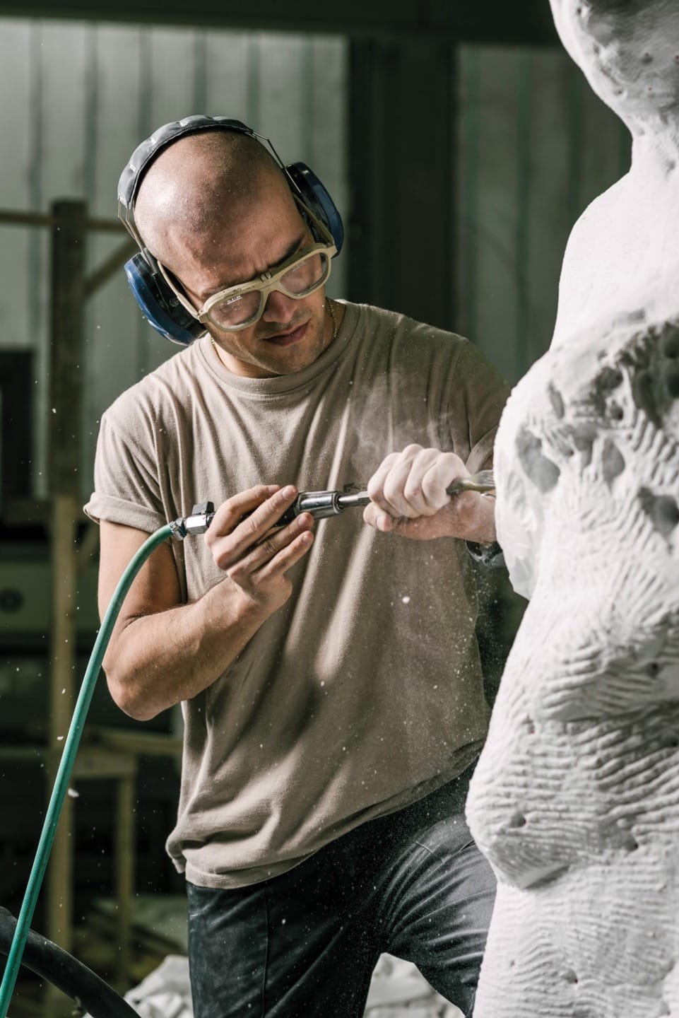 Sculptor wearing ear protection and safety glasses working on a marble statue with a pneumatic tool in a studio.