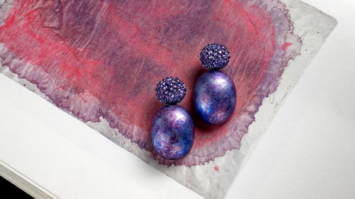 Purple gemstone earrings with textured orbs displayed on a watercolor paper background with red and purple hues.