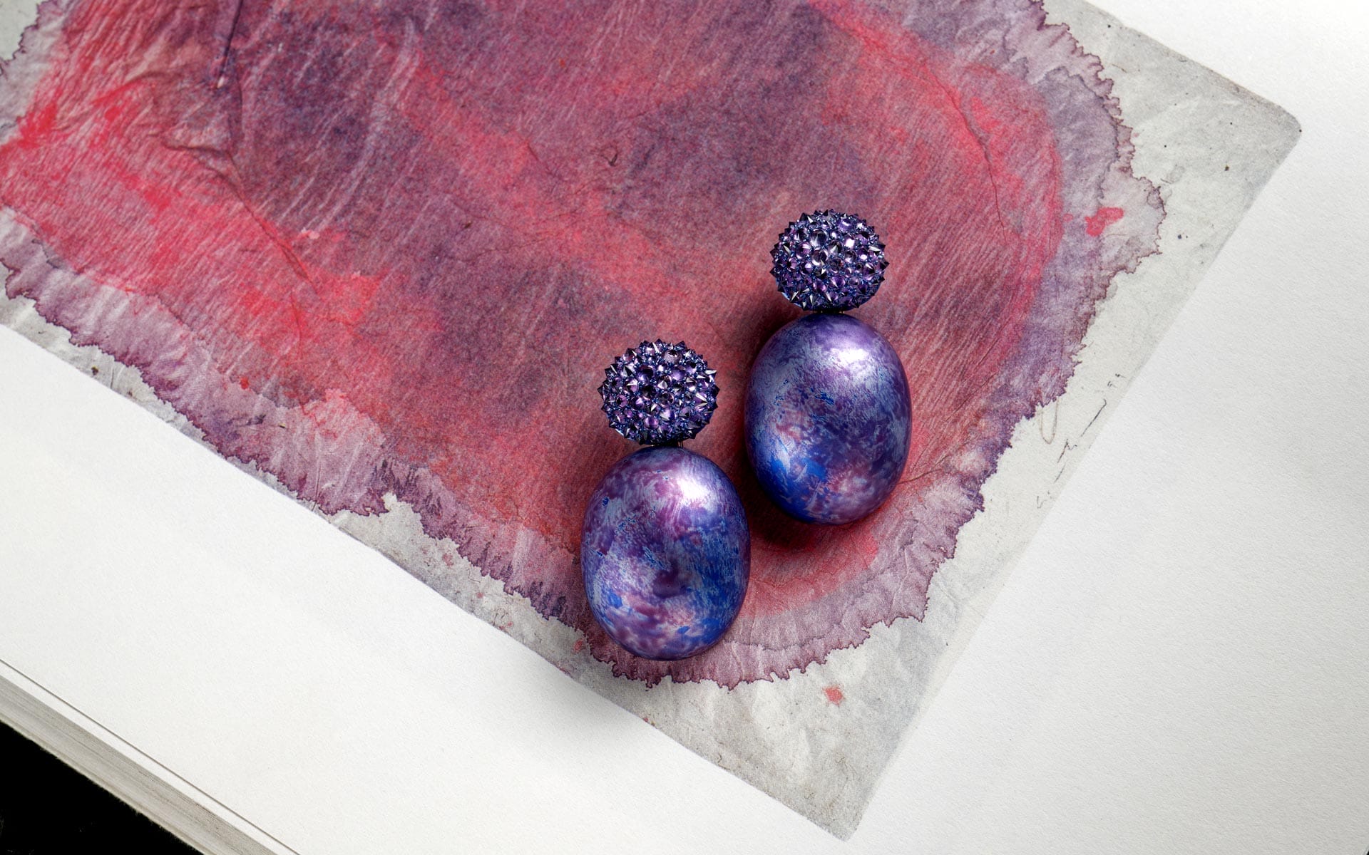 Purple gemstone earrings with textured orbs displayed on a watercolor paper background with red and purple hues.