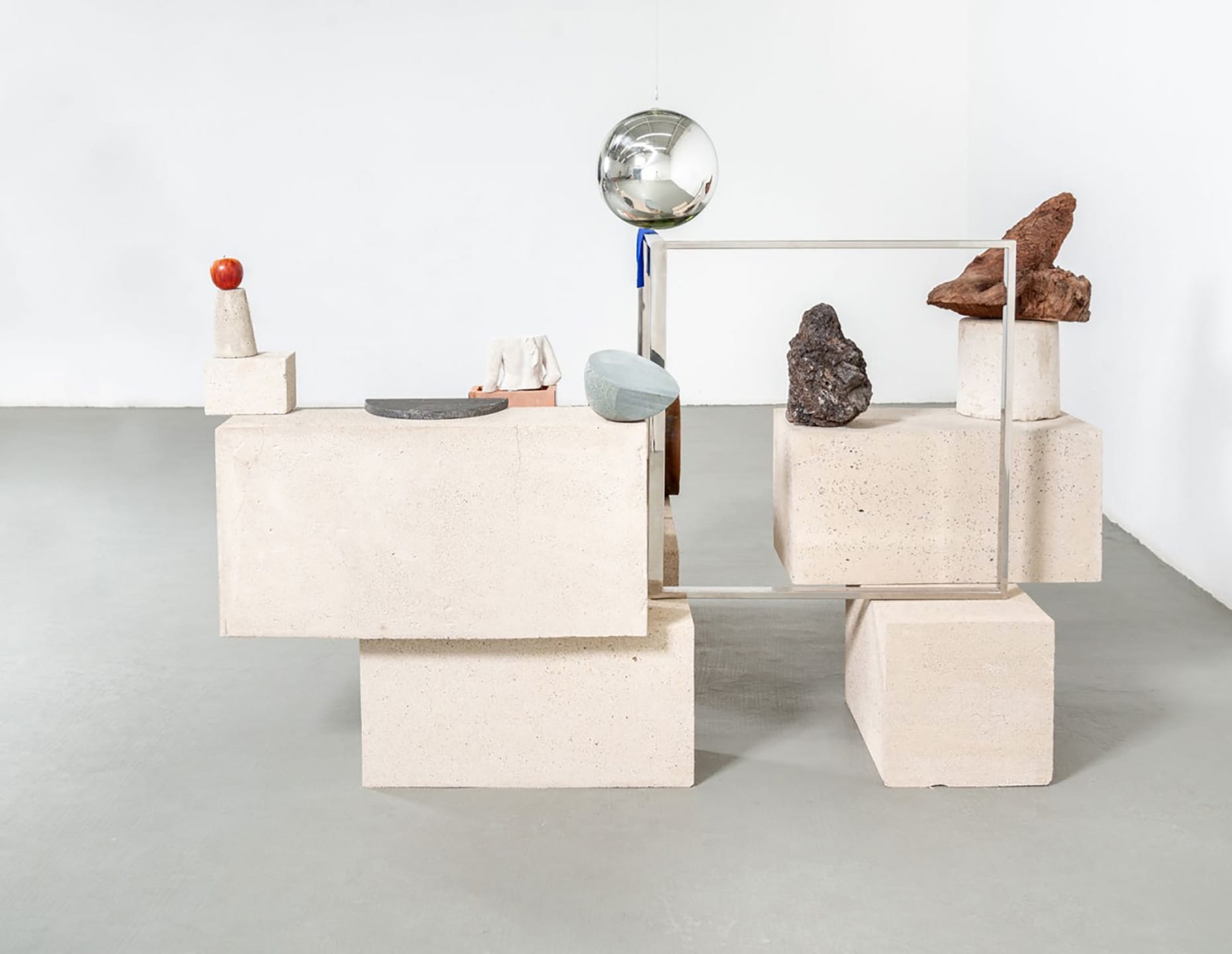 Contemporary sculpture with geometric shapes, stones, a sphere, and a red sphere on a white floor and wall background.