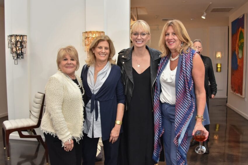 Group of four women standing together, smiling, in a modern art gallery setting with colorful paintings in the background.