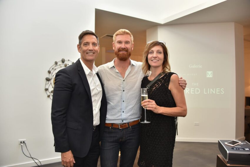 Three people at an art gallery event, two men and a woman with a drink, standing in front of a modern white backdrop.