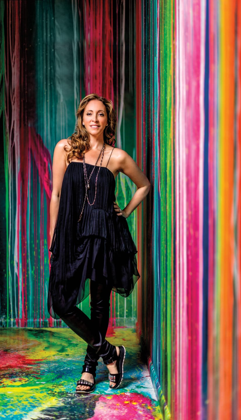Woman standing in a colorful, artistic hallway wearing a black dress and sandals, with vibrant paint streaks in the background.