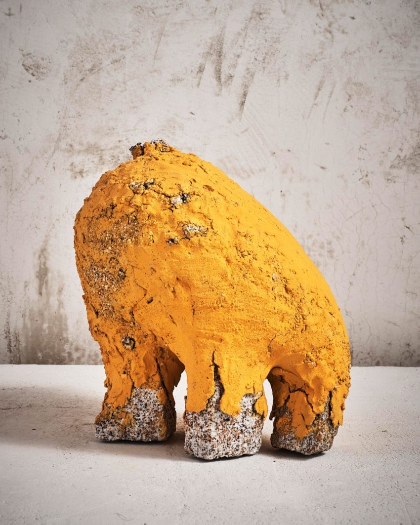 Abstract orange and gray textured sculpture with a rounded shape against a neutral, textured wall background.