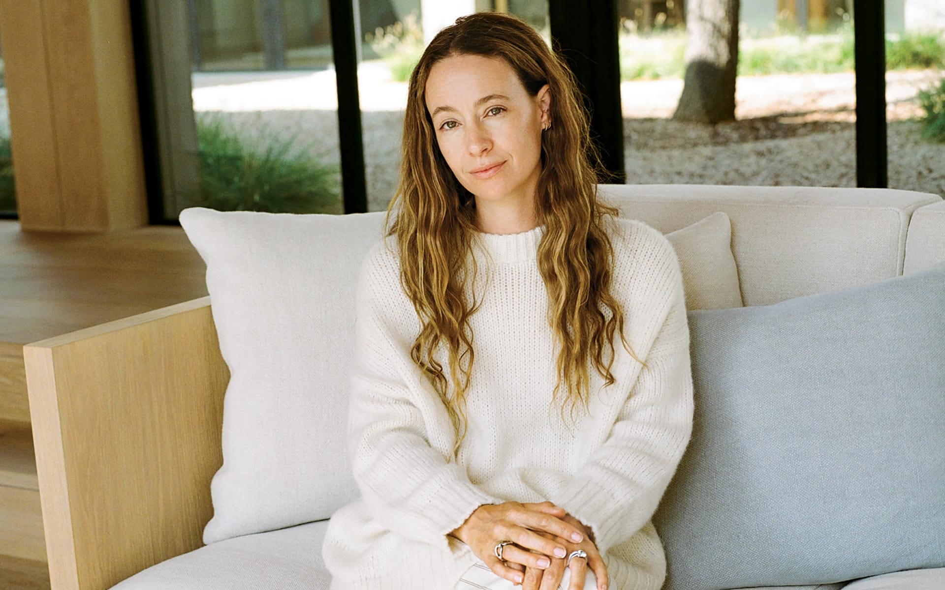 Person sitting on a couch wearing a white sweater, with a relaxed expression and long hair, in a modern living room.