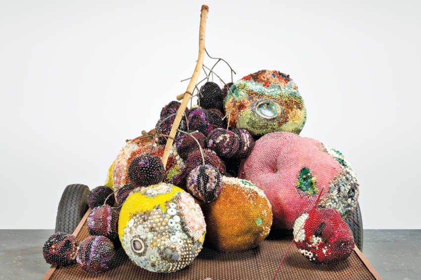 Colorful beaded sculptures resembling round fruits with a branch, displayed on a textured surface against a plain background.
