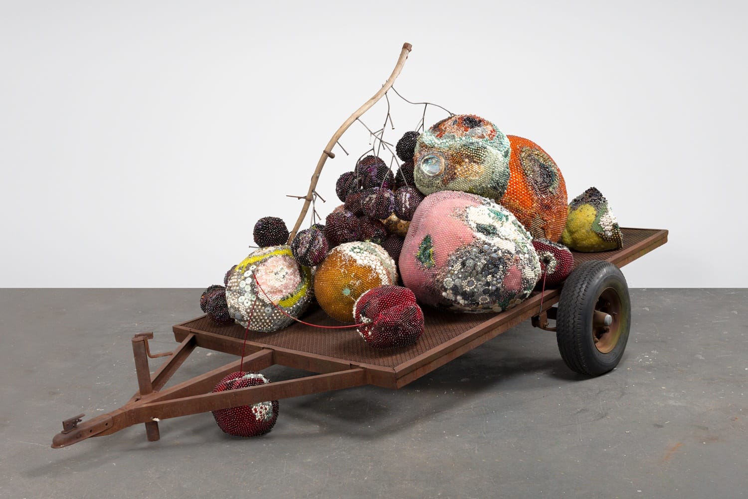 Colorful abstract sculpture of various textured spheres on a rusty metal cart with wheels in a minimalist gallery setting.