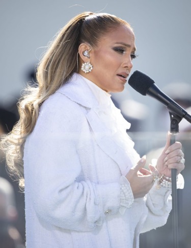 Singer performing on stage, wearing a white outfit and earrings, holding a microphone, long hair styled in a ponytail