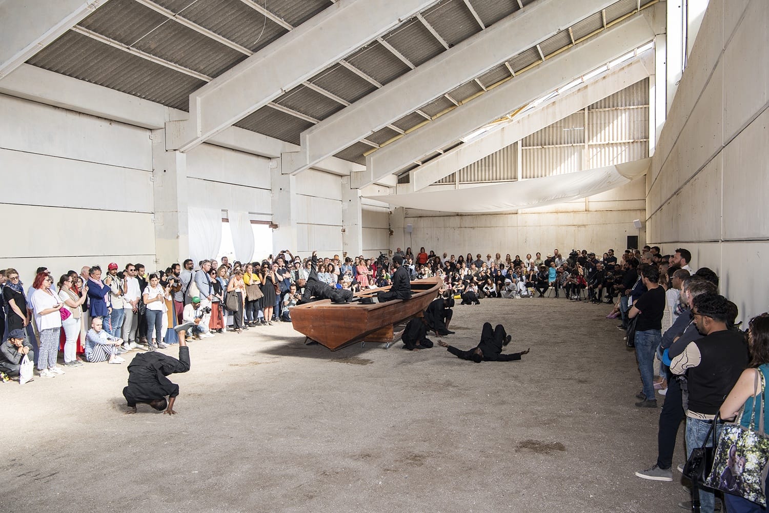 Audience watching a theatrical performance inside a large industrial building with performers around a wooden boat on the floor