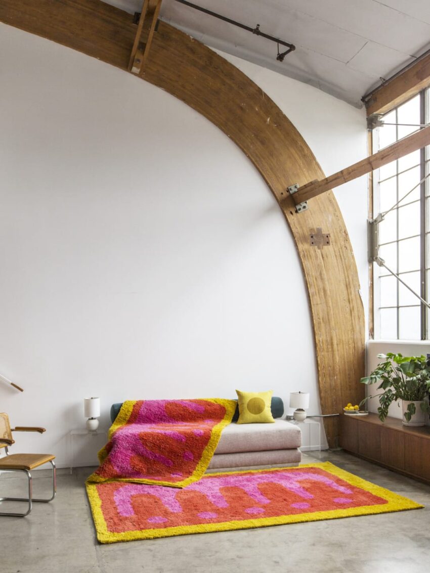 Colorful modern living room with a pink and yellow textured rug and blanket, arched wooden beams, and large windows.