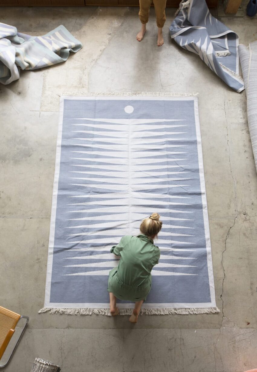 Person in green shirt inspecting a blue and white patterned rug on a concrete floor with other rolled-up rugs nearby