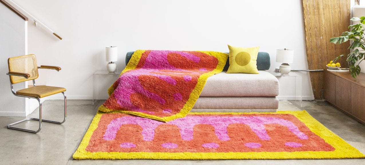 Colorful modern living room with patterned pink and yellow rug, matching blanket on beige sofa, and a wooden chair.