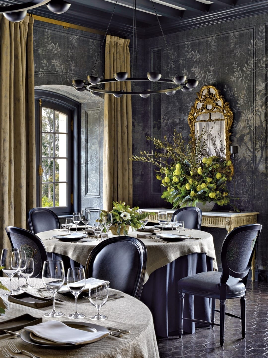 Elegant dining room with dark walls, round tables set with white tableware, floral centerpieces, and an ornate mirror.