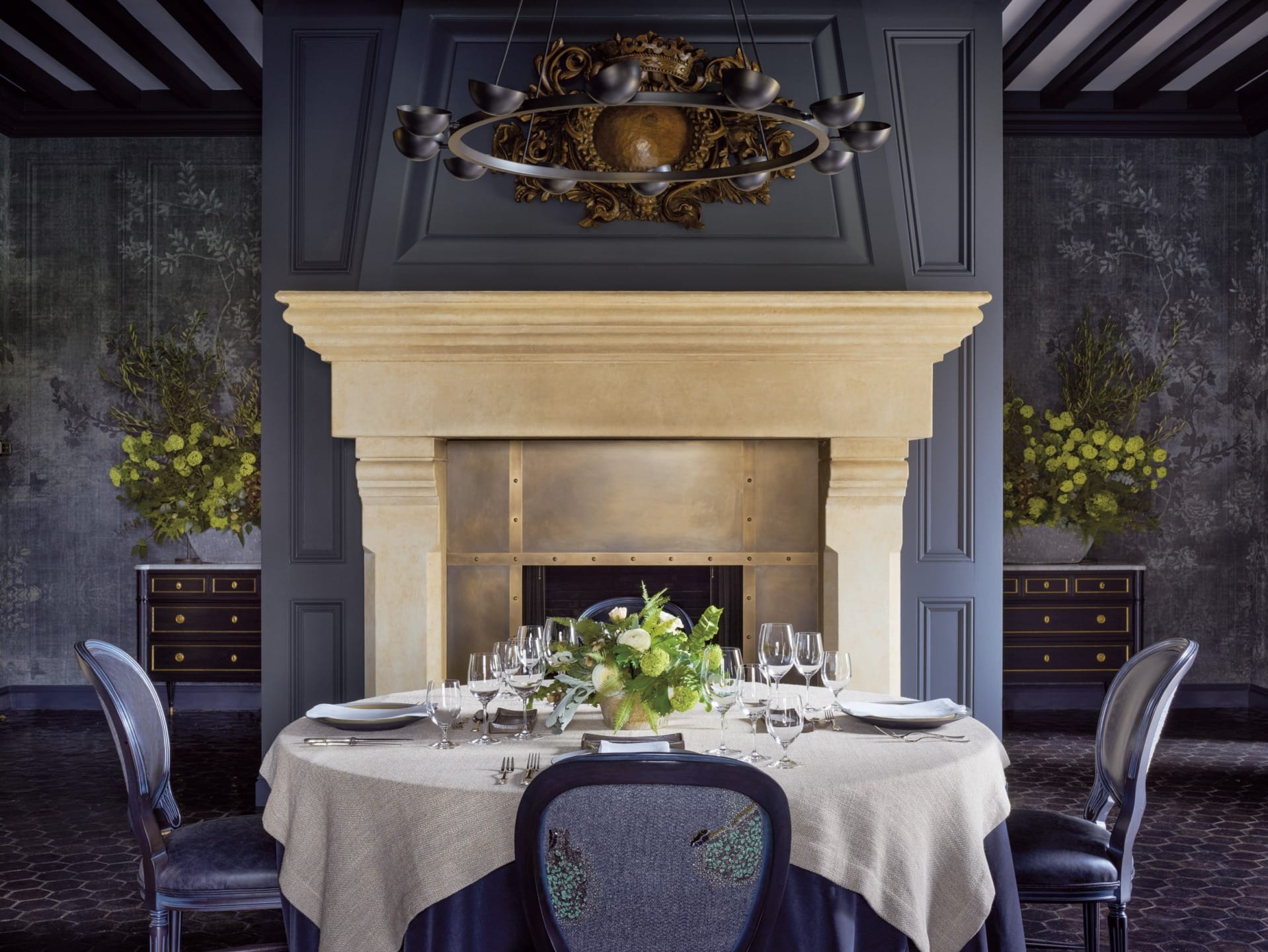 Elegant dining room with a round table, beige tablecloth, floral centerpiece, and a classic stone fireplace in the background.