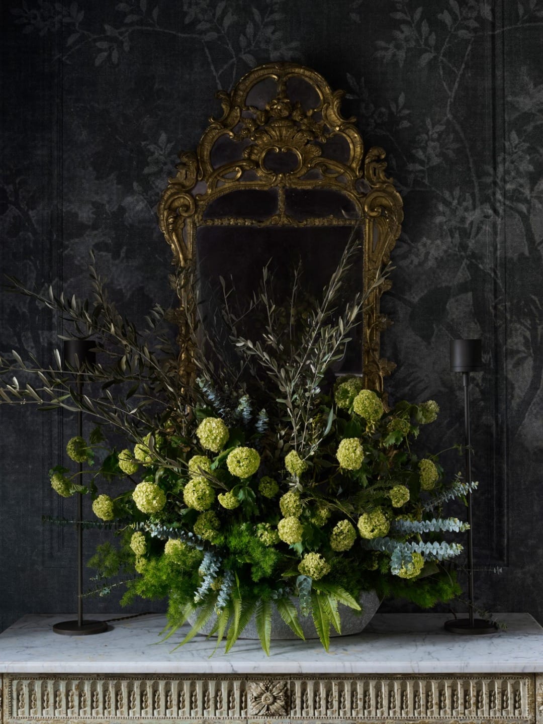 Ornate mirror with lush green floral arrangement and candles on a marble table against a dark floral wallpaper.