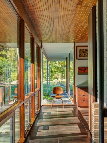 A modern hallway with large windows, wood ceiling, and a chair overlooking a green outdoor scene.