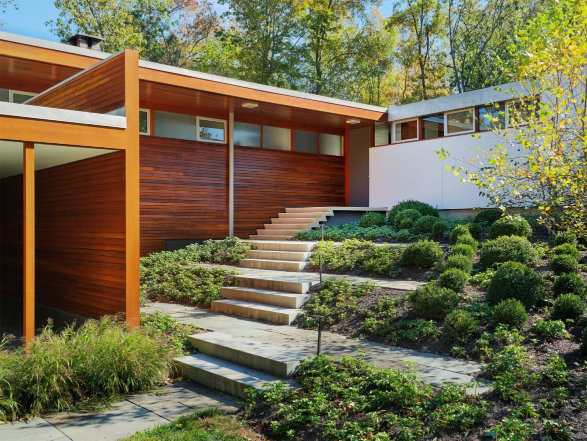Mid-century modern house with wooden facade, large windows, and landscaped garden featuring trimmed bushes and a stone pathway.