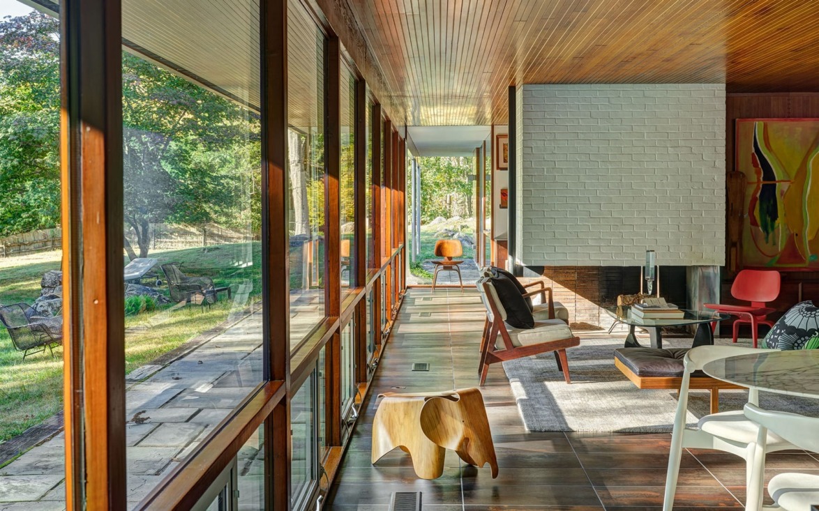 Modern living room with wooden ceiling, large windows, and cozy seating area overlooking a green garden with stone pathway.