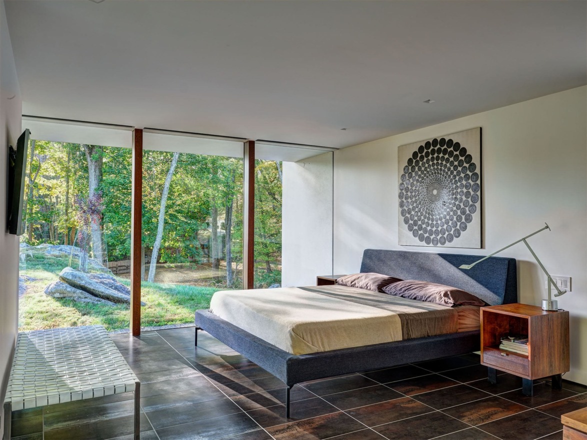 Modern bedroom with large glass windows overlooking a forest, featuring minimalist furniture and a geometric wall art.