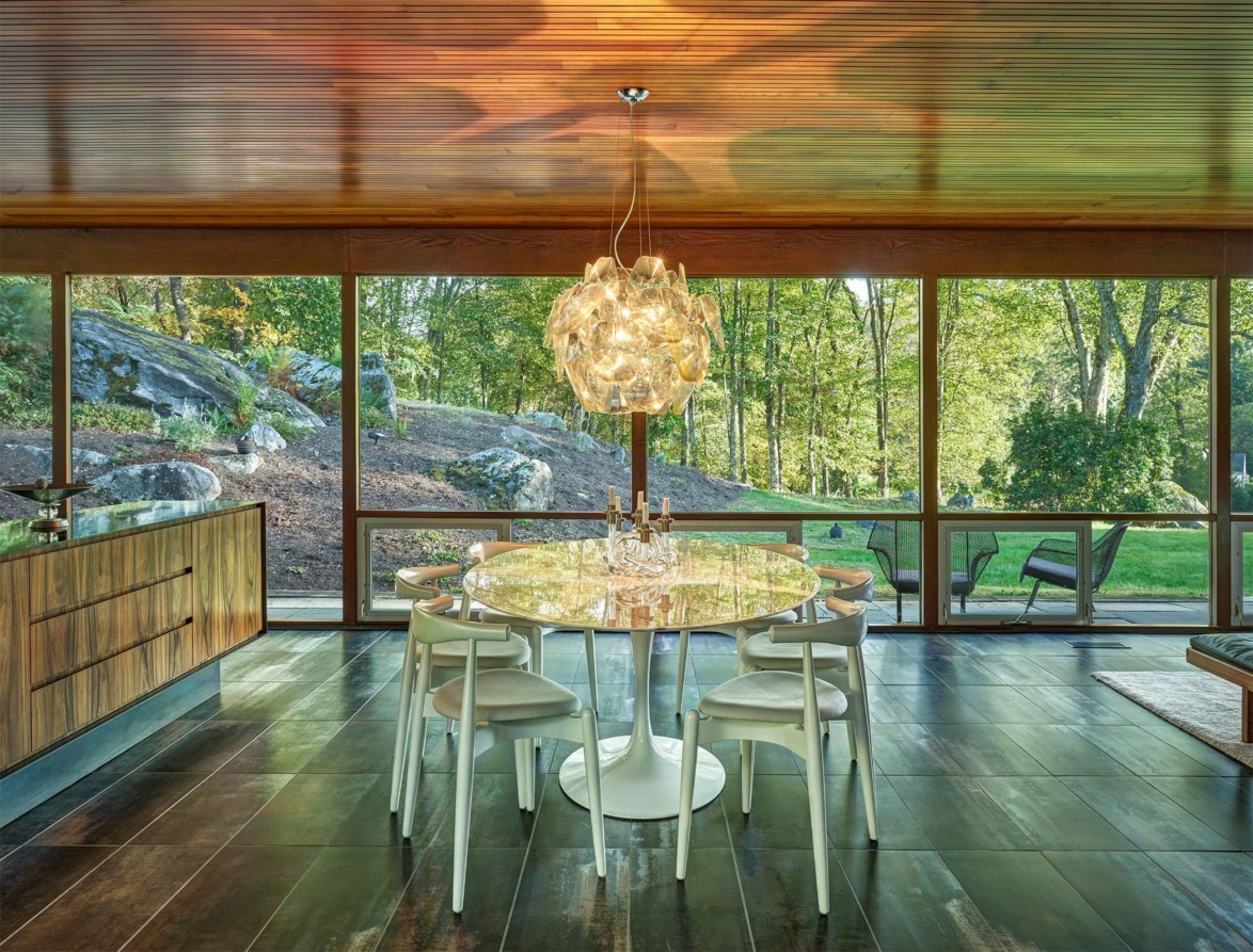 Modern dining room with glass walls, round table, unique chandelier, and view of lush outdoor garden.